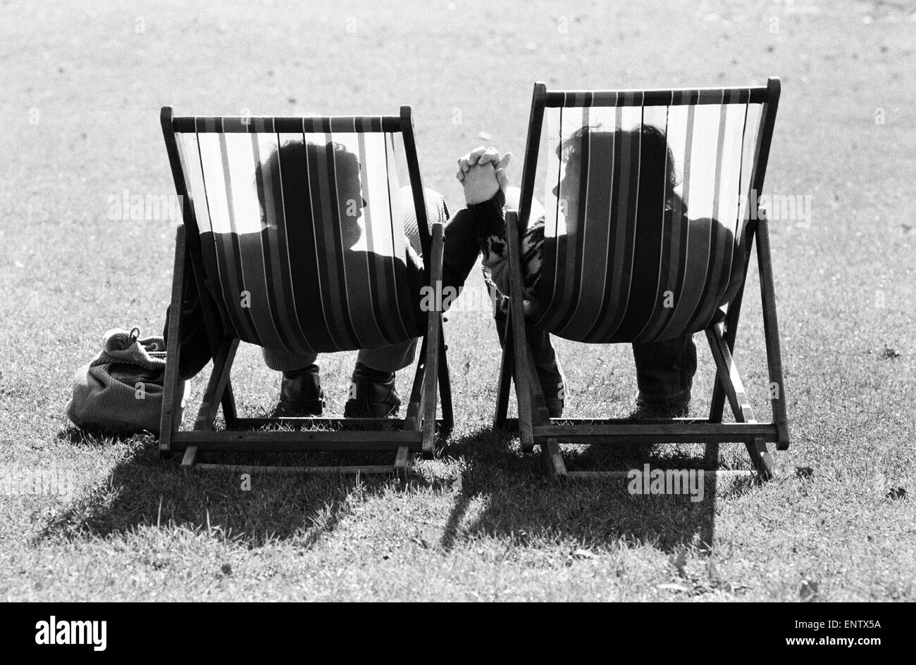A couple in sit in a pair of deckchairs holding hands and looking at one another as they enjoy the warm spring sunshine. 4th March 1979. Stock Photo