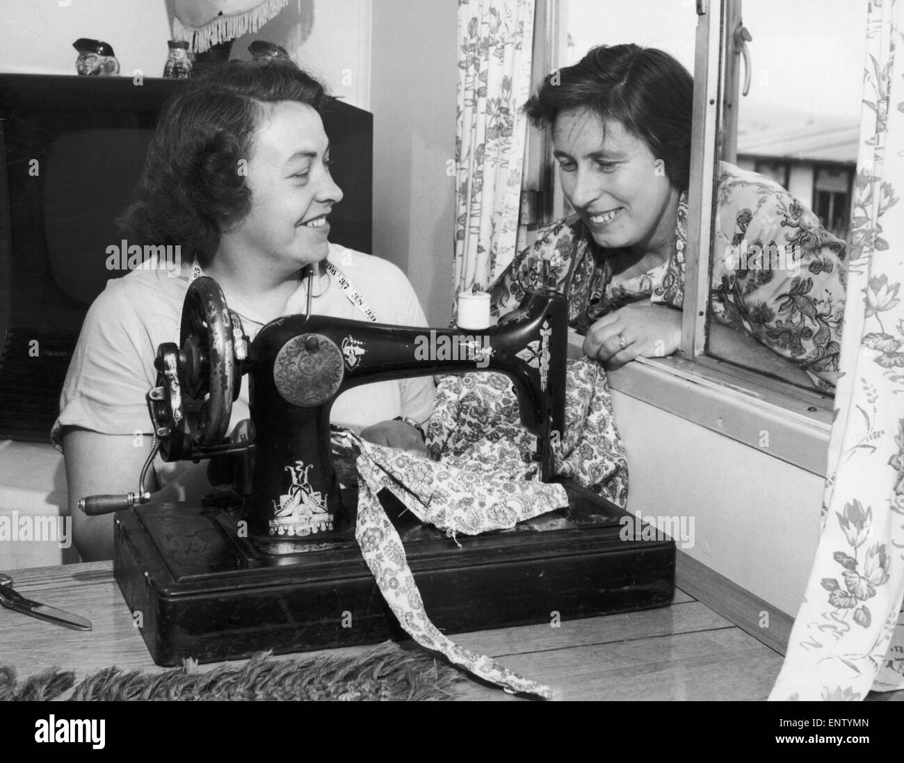 Busy at her sewing machine is Mrs. Beaty Evans, her next door neighbour, Mrs. Betty Davies looks in at the window to discuss the latest bathing costume. July 1955 Stock Photo