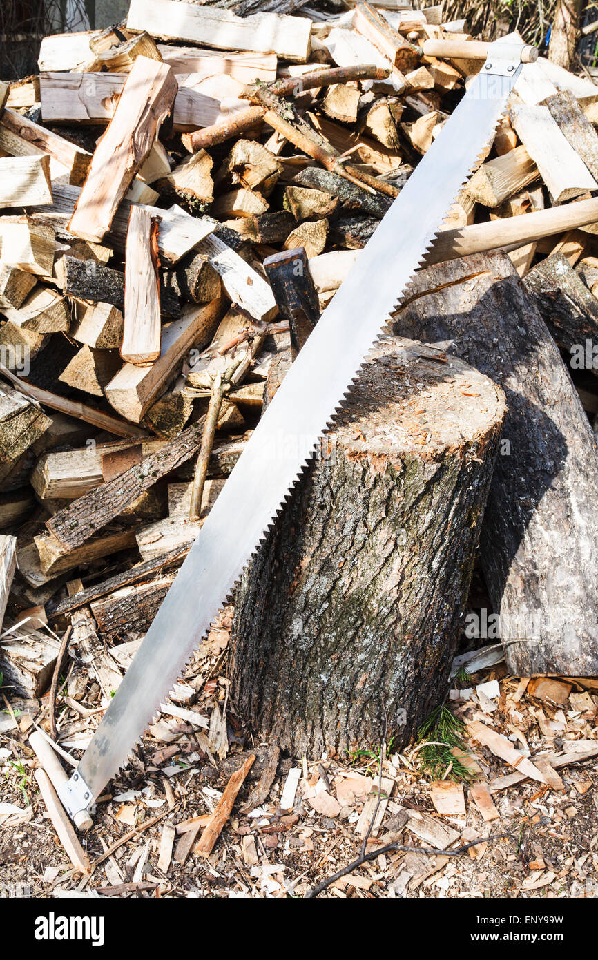 two-handled saw and ax in deck for chopping firewood, pile of wood on rustic courtyard Stock Photo