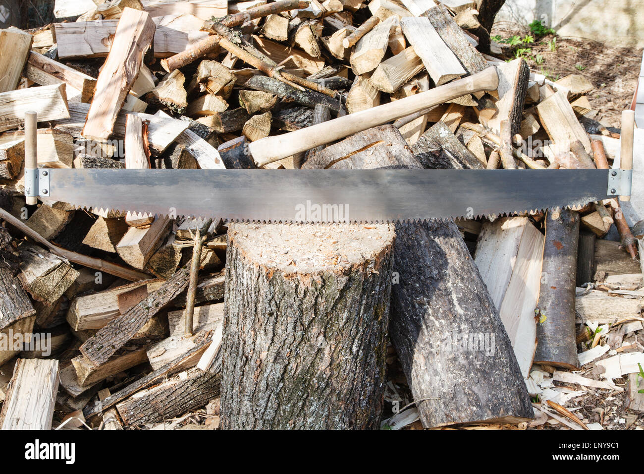 two-handled saw and ax in chopping block, pile of firewood on rustic courtyard Stock Photo