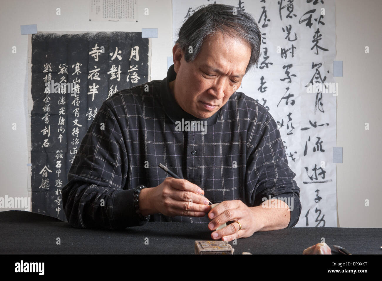 Asian man carving Chinese seal stamp in Gaithersburg, Maryland. Calligraphy panels can be seen in the background. Stock Photo