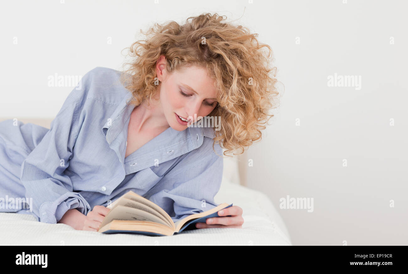 blonde woman reading a book while lying on her bed Stock Photo