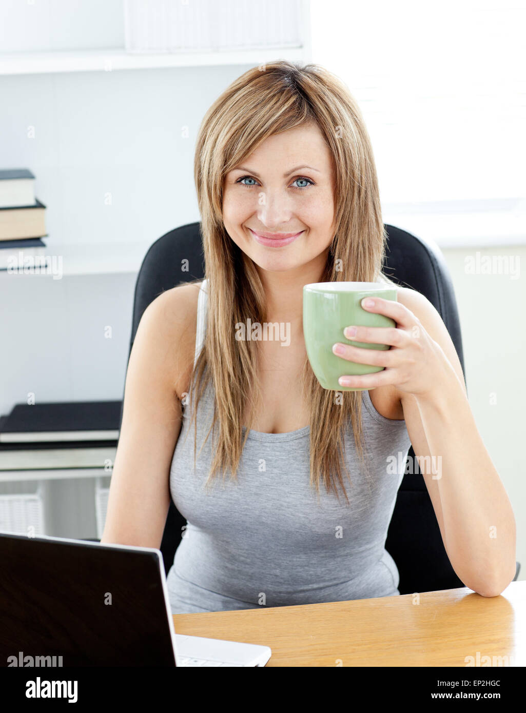 Glowing businesswoman holding a cup of coffee using her laptop at her ...