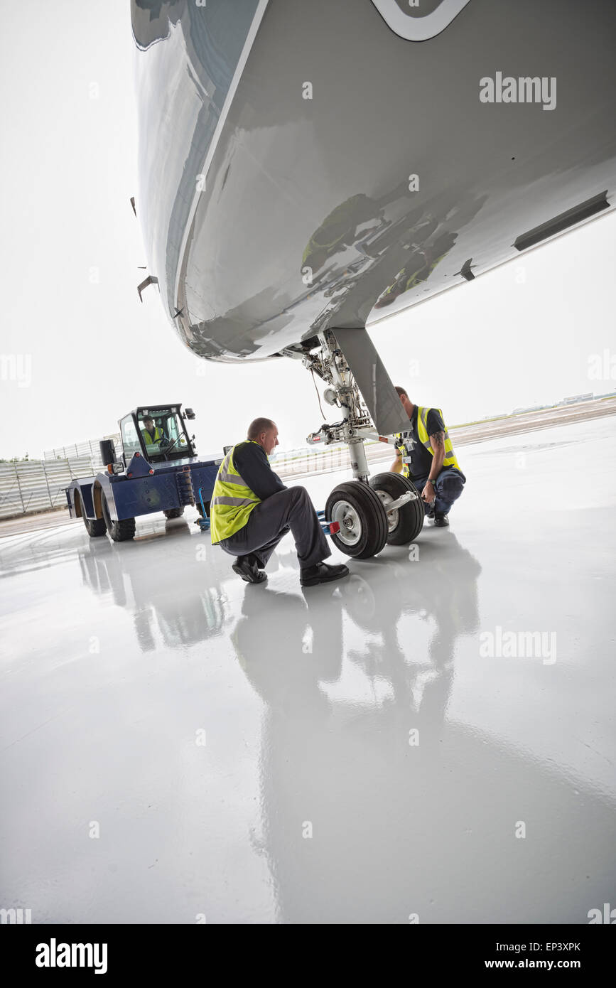 Ground crew attaching a tow bar to an aircraft's nose landing gear with a towing tractor ready to pushback the airplane Stock Photo