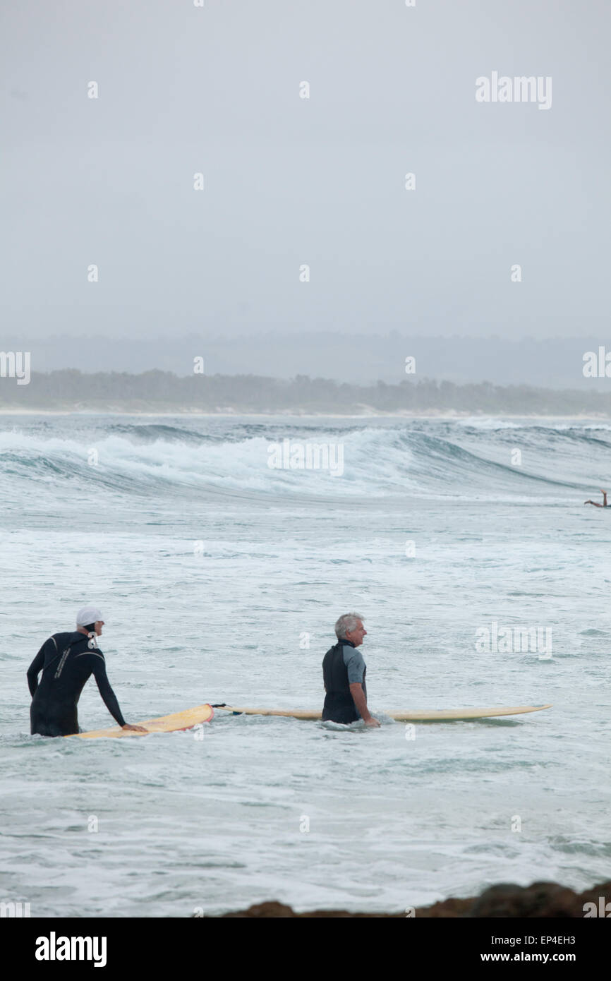 Two surfers stand in the water gazing at the surf rolling by in Byron Bay, Australia. Stock Photo