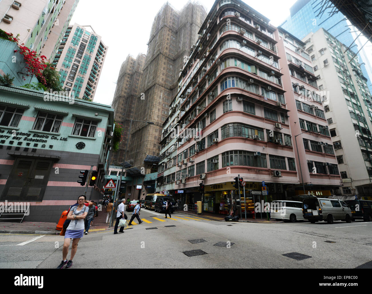 A street corner in Hong Kong featuring old residential buildings with curved balconies, under construction skyscrapers covered in scaffolding, and a woman standing at the crosswalk while others walk by.