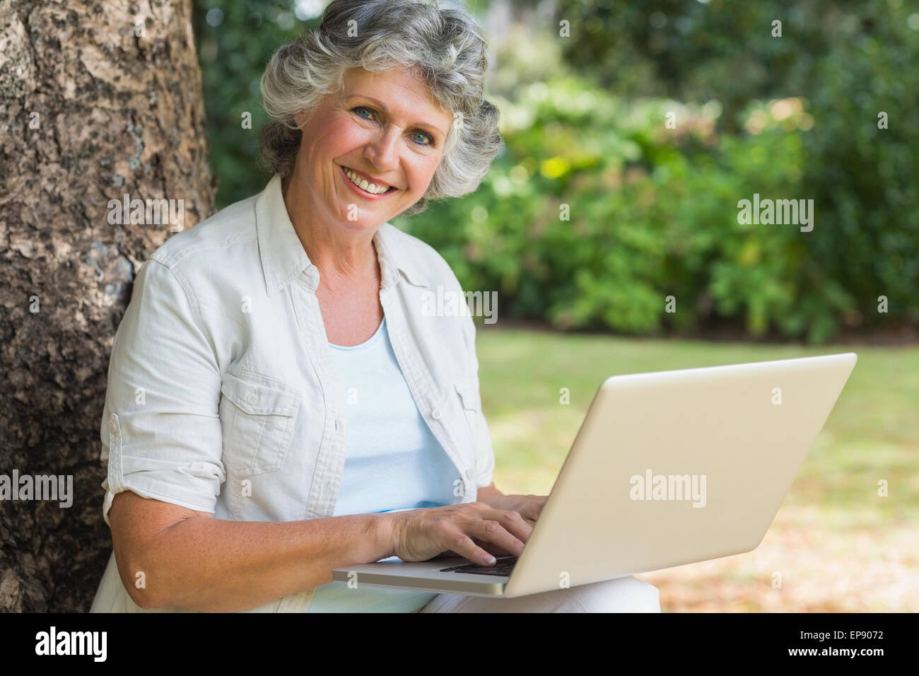 Cheerful mature woman using a laptop sitting on tree trunk Stock Photo