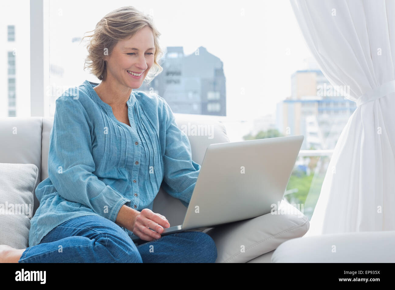 Cheerful blonde woman sitting on her couch using laptop Stock Photo - Alamy