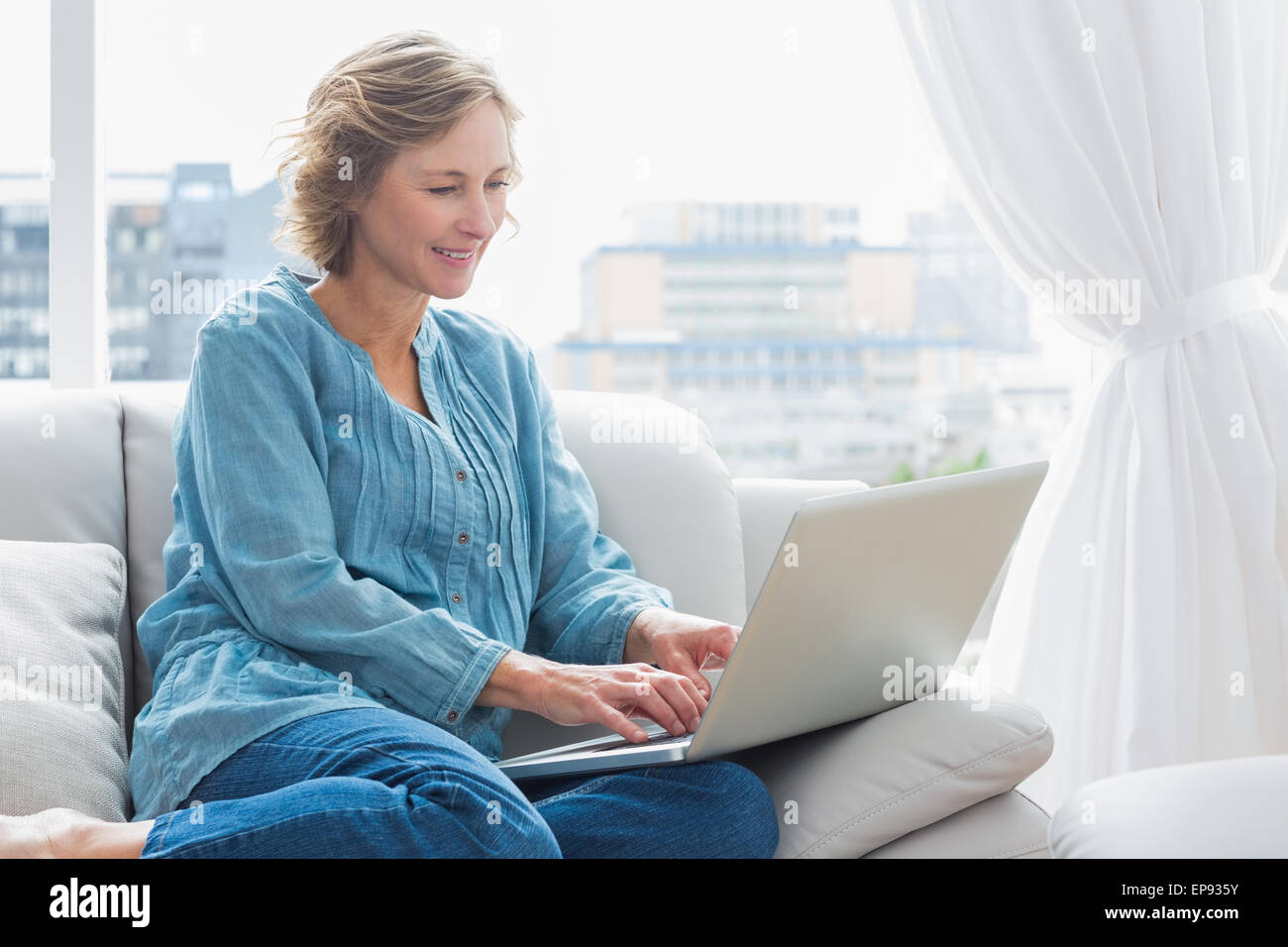 Happy blonde woman sitting on her couch using laptop Stock Photo - Alamy