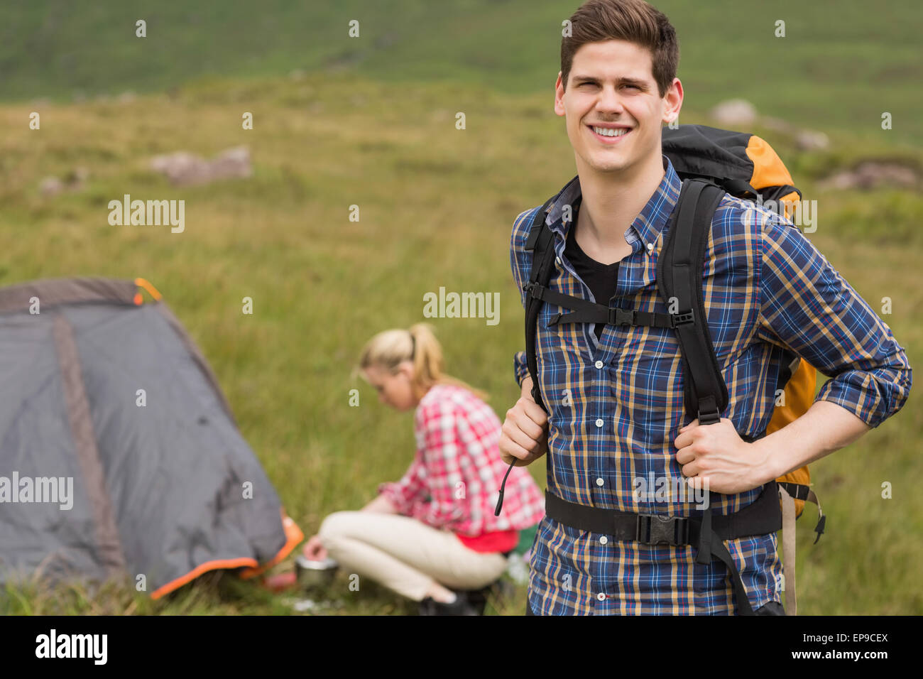 Cheerful man carrying backpack while girlfriend is pitching tent Stock Photo