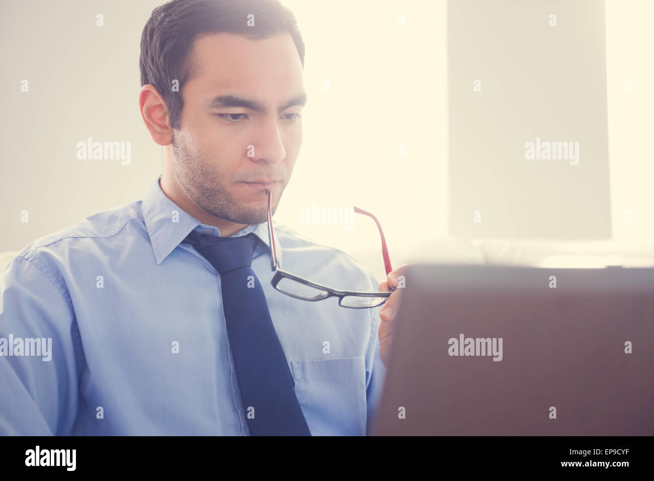 Unsmiling man biting his eyeglasses and using a laptop Stock Photo