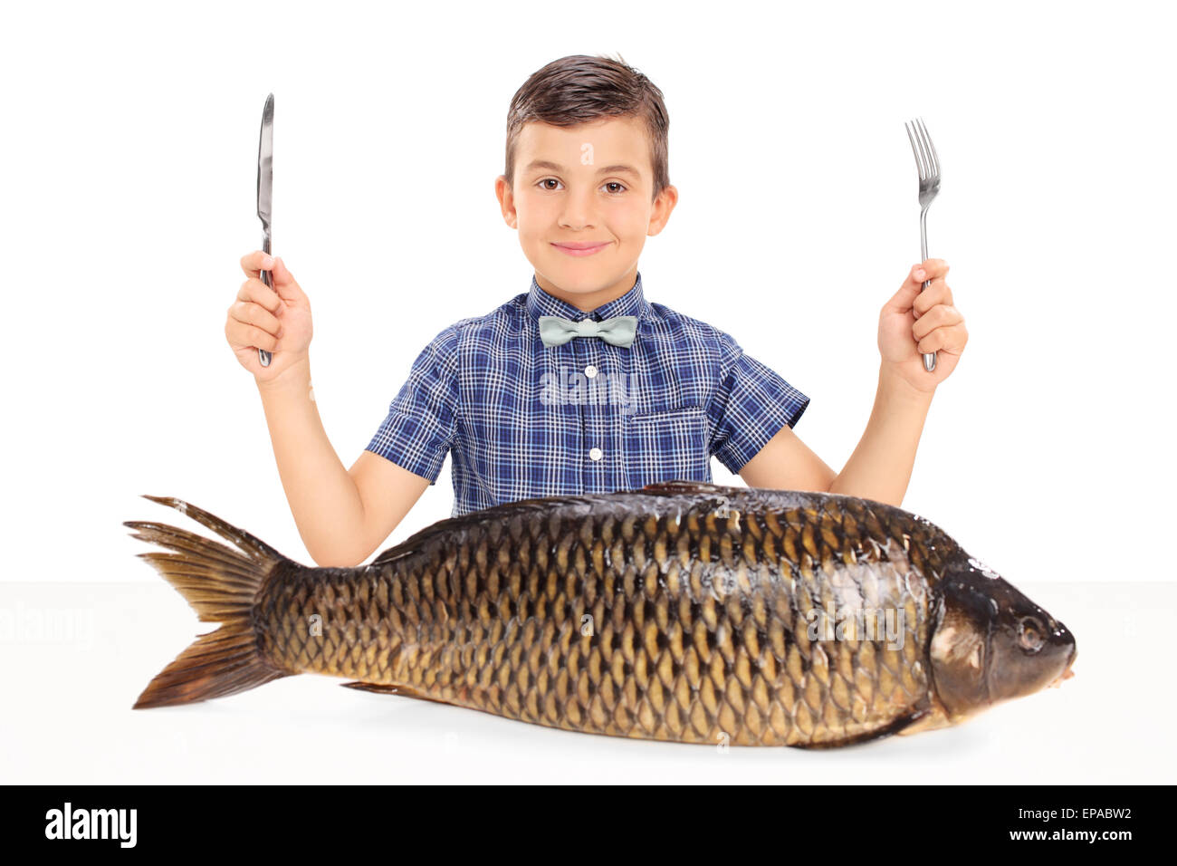 Little kid holding a fork and a knife seated at a table with a huge raw fish in front of him isolated on white background Stock Photo