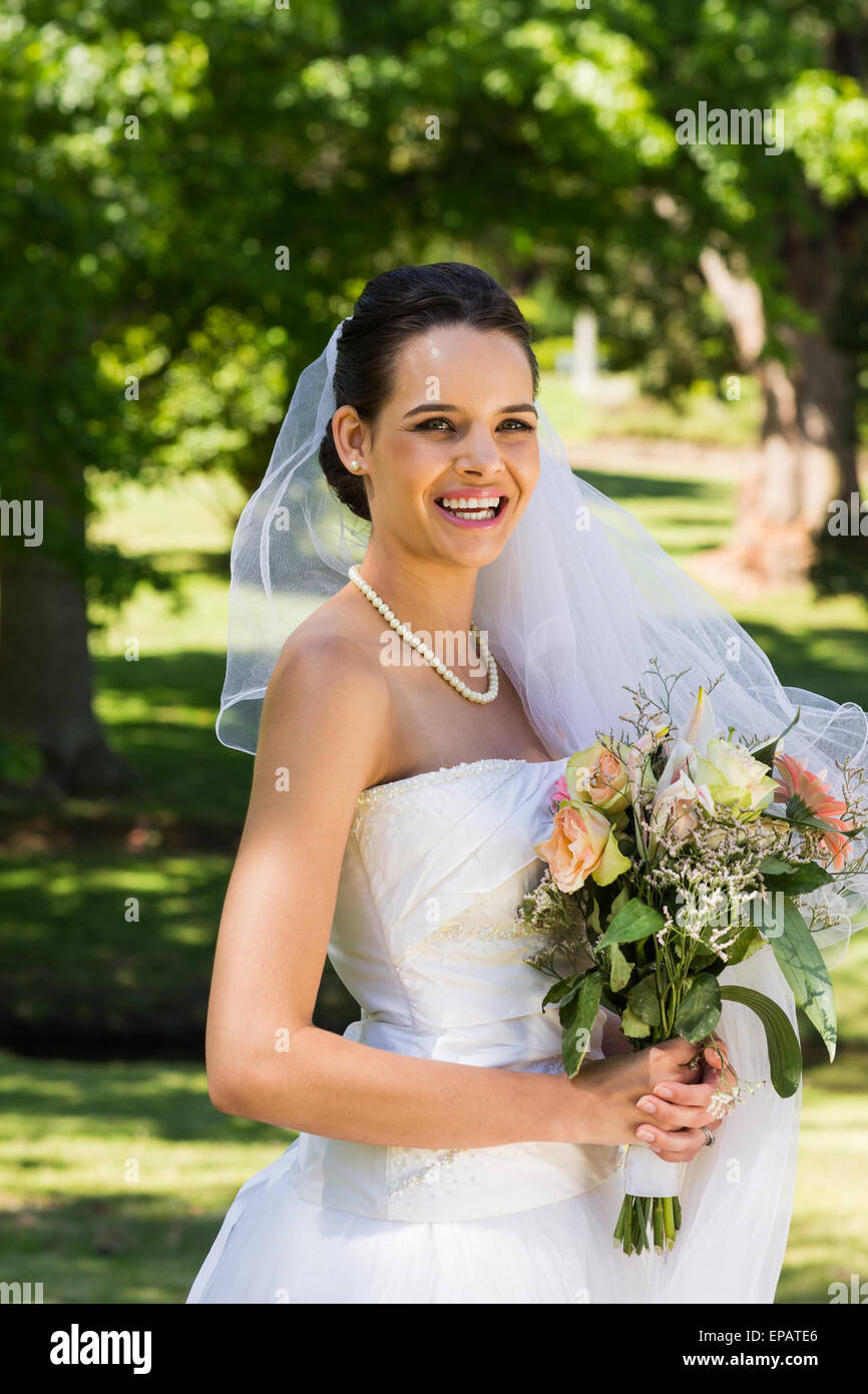 Smiling beautiful bride with bouquet standing in park Stock Photo - Alamy