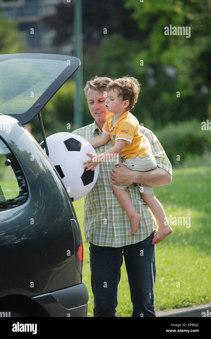 Father holding son on his arm standing at the back of his car, son is grabbing an air ball Stock Photo