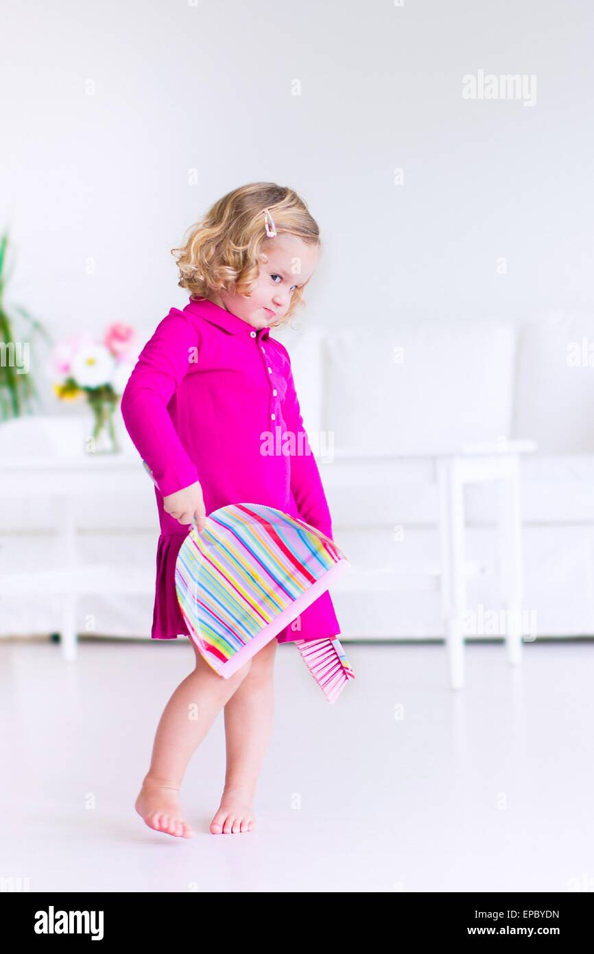Funny curly little girl in a pink dress helping at home cleaning the floor sweeping in a white sunny living room Stock Photo