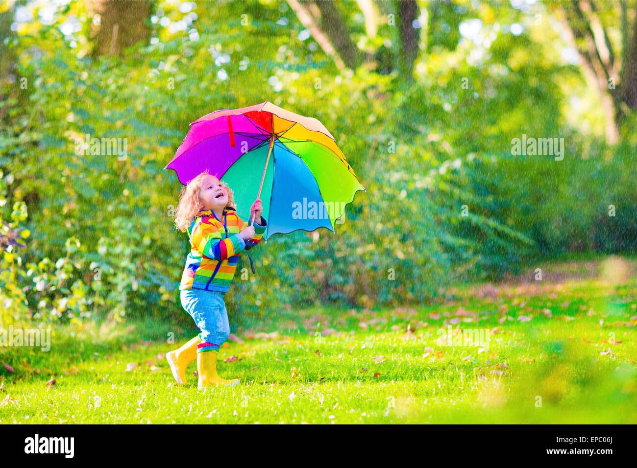 Funny cute curly toddler girl wearing yellow waterproof coat and boots holding colorful umbrella playing in the garden by rain Stock Photo