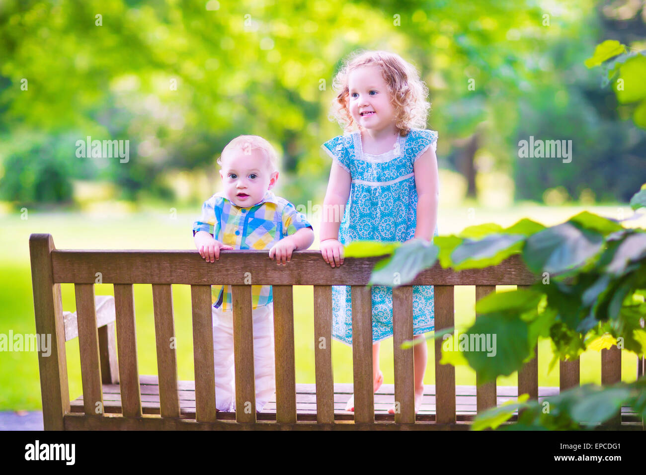 Adorable kids, little curly girl and a cute baby boy, brother and sister, sitting together on a wooden bench in a garden Stock Photo