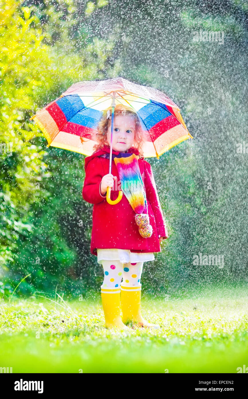 Funny cute curly toddler girl holding colorful umbrella playing in the garden by rain and sun weather on a warm autumn or summer Stock Photo