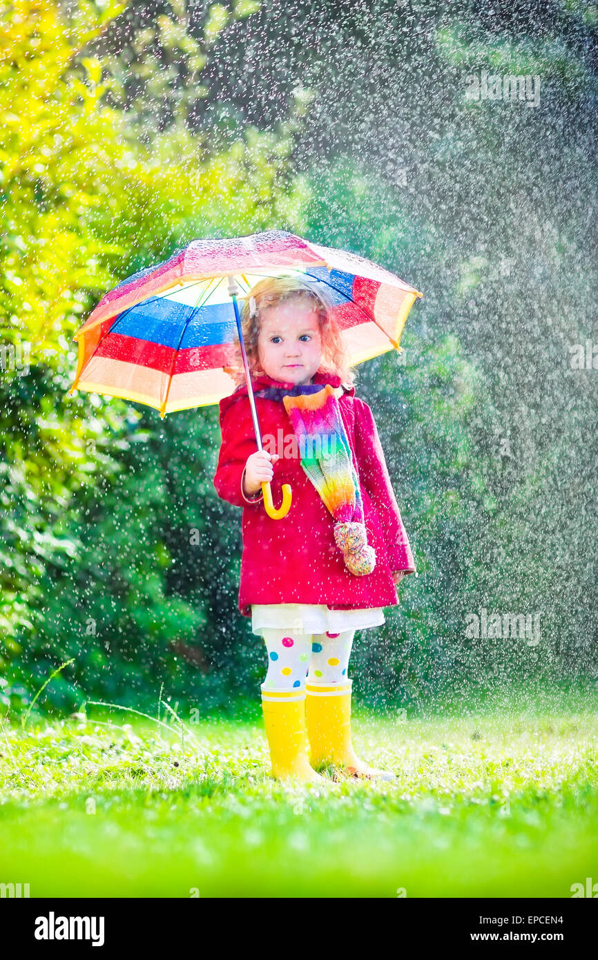 Funny cute curly toddler girl holding colorful umbrella playing in the garden by rain and sun weather on a warm autumn or summer Stock Photo