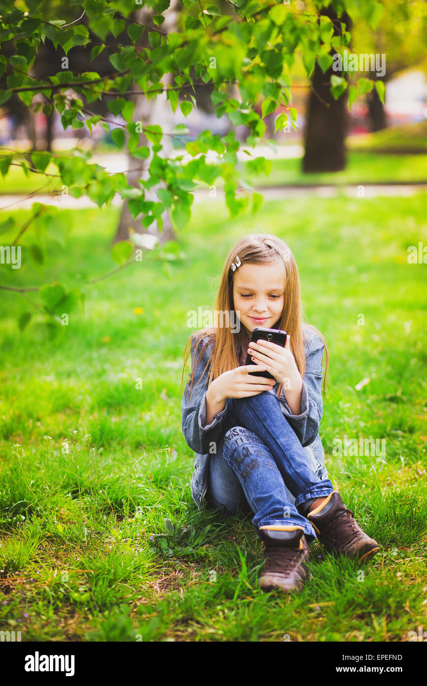 portrait of smiling teenage girl sitting in park and texting sms using mobile phone Stock Photo