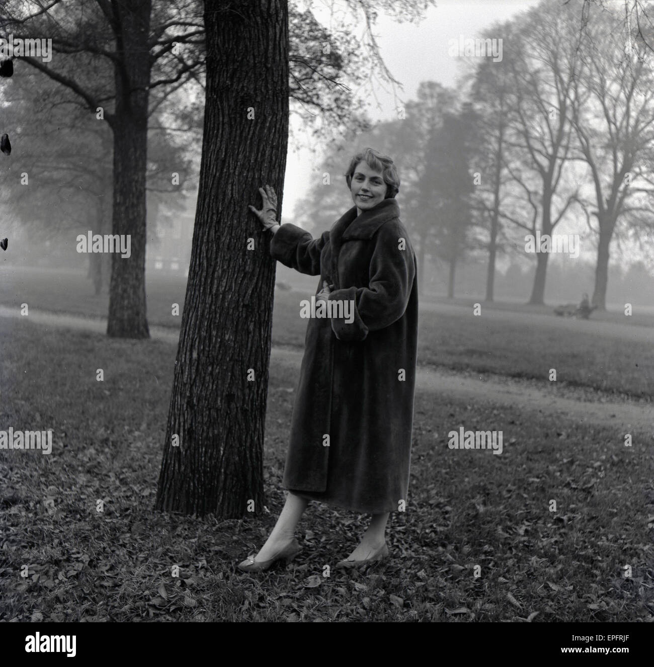 1950s, historical, a lady wearing a long fur coat stands beside a tree in a park. Stock Photo