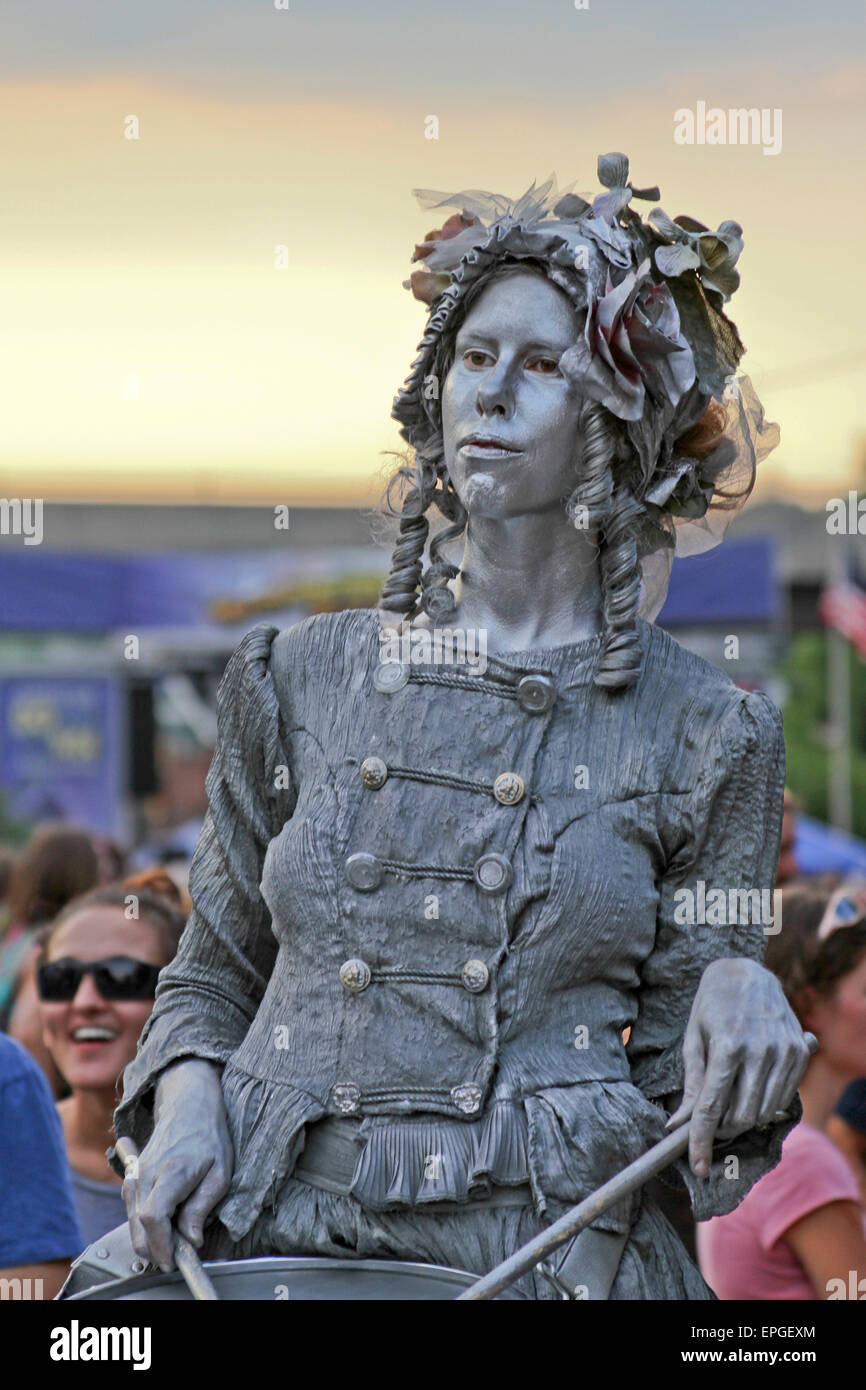 Close up of a young woman coated in silver and playing a drum as she performs as a living statue for tips Stock Photo
