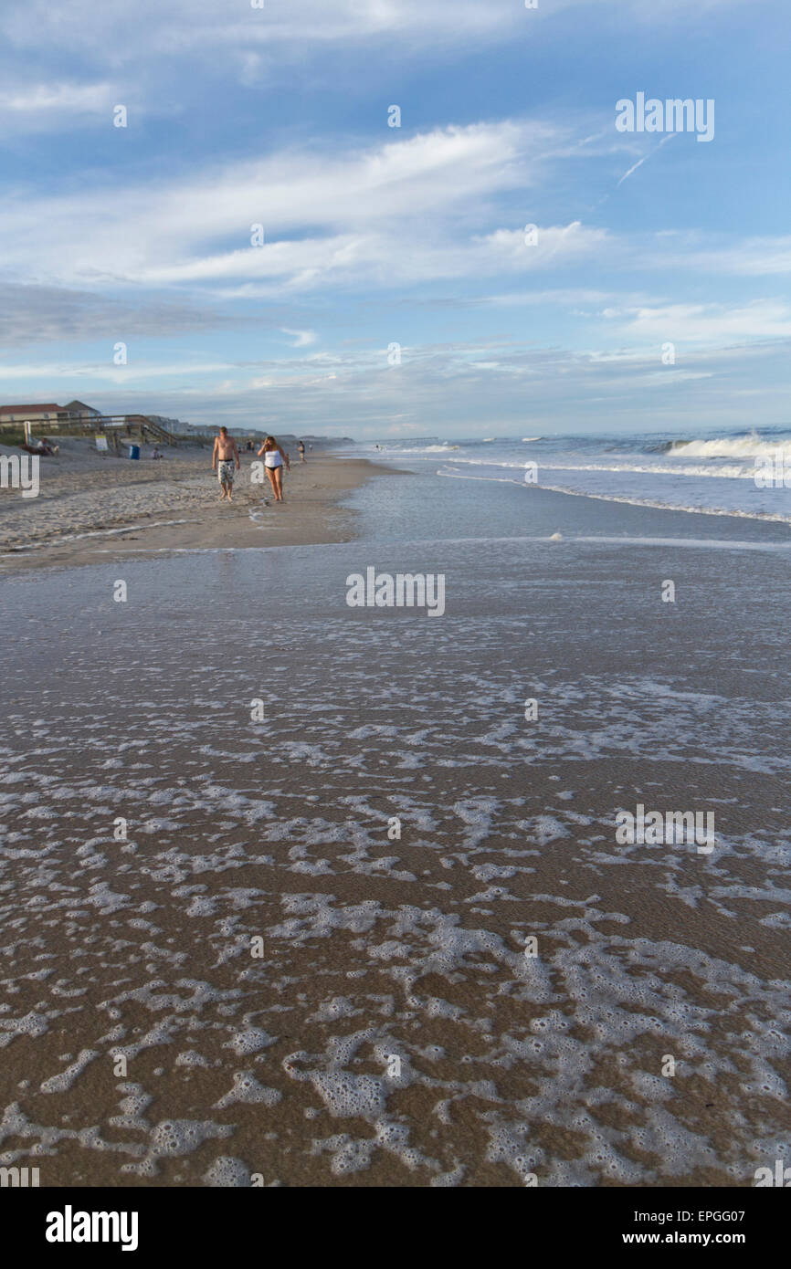 The foamy tide comes in covering the beach in front of people walking along the ocean under blue sky and bright puffy clouds Stock Photo