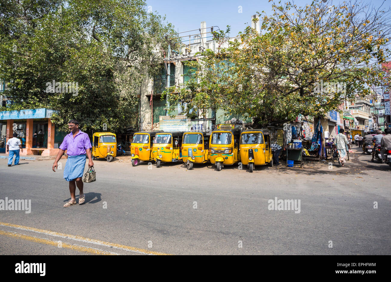 Local Indian man crossing the road in front of a row of parked yellow auto rickshaws taxi cabs, street scene, Chennai, Tamil Nadu, India Stock Photo