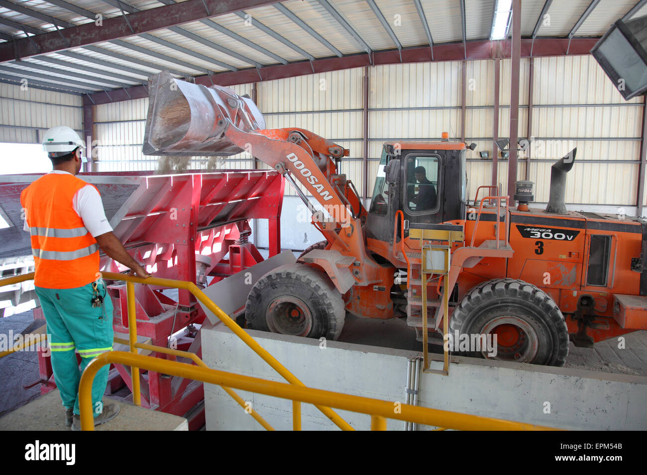 A large excavator tips waste material into a hopper at a large aluminium smeltting plant watched by a supervisor Stock Photo