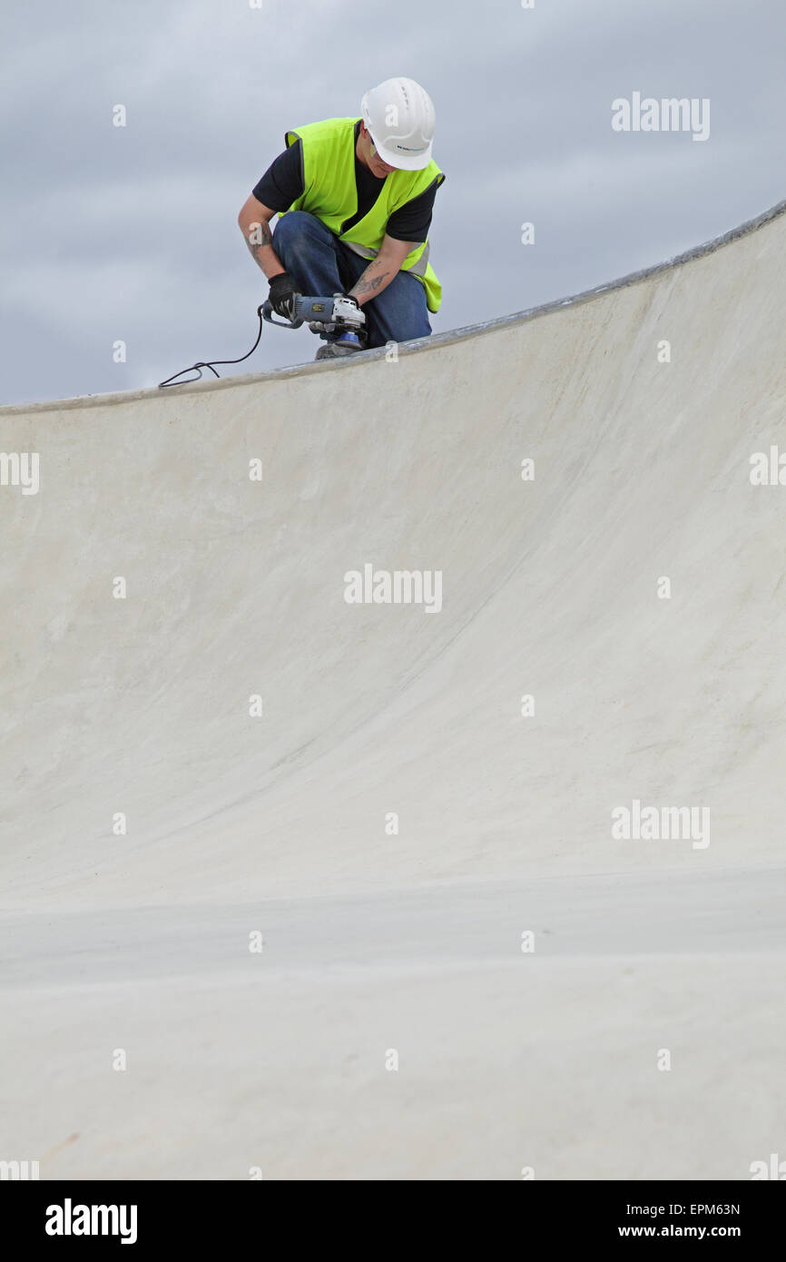 A construction worker uses a grinding tool to finish the rim of a concrete bowl at a skateboard and BMX bike park. Stock Photo