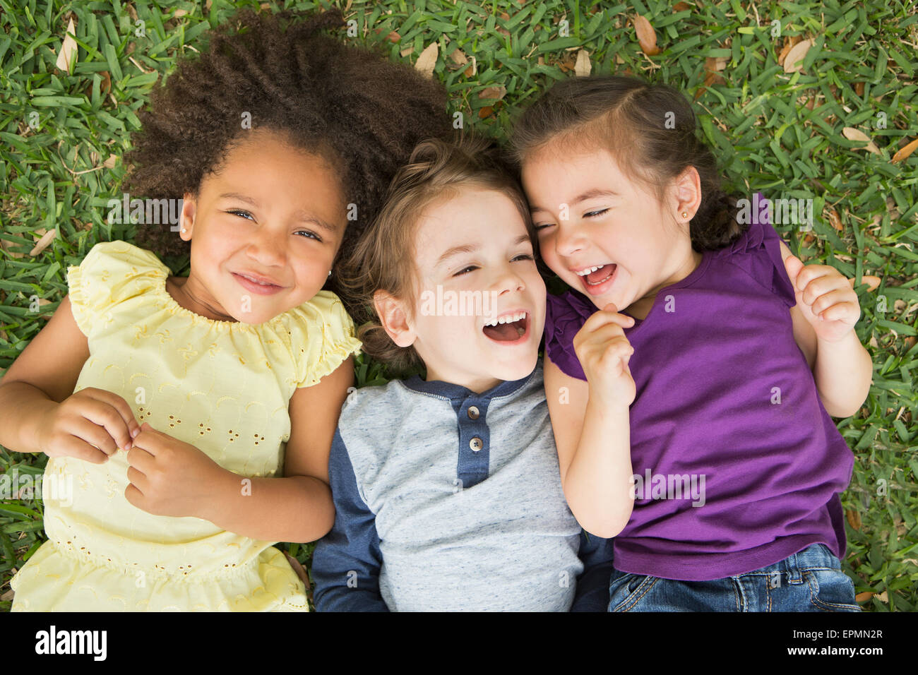 Three children lying on their backs on the grass, looking up and laughing. Stock Photo
