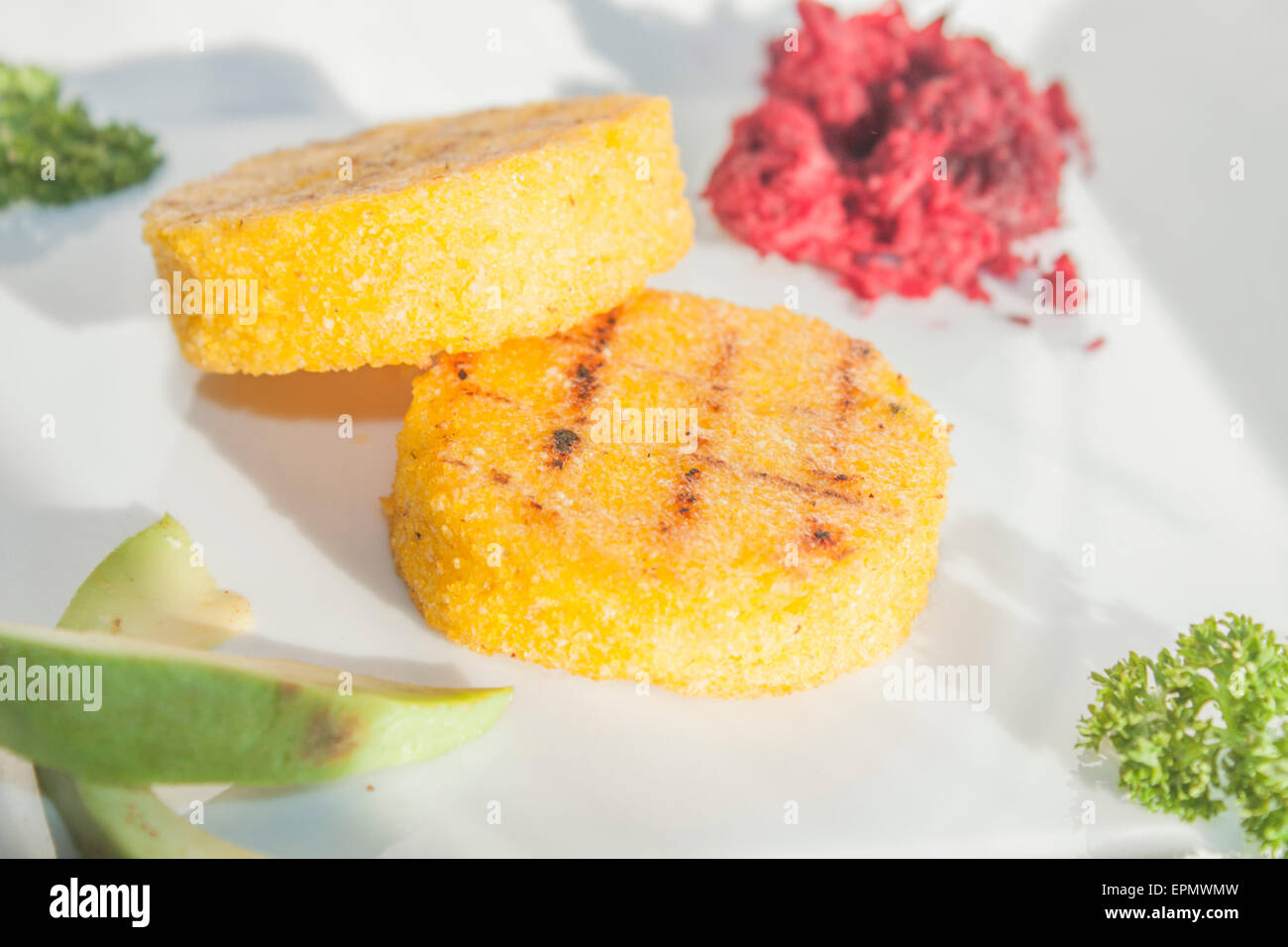 Two yellow polenta slices, near avocado, parsley and beetroot, on a white table Stock Photo