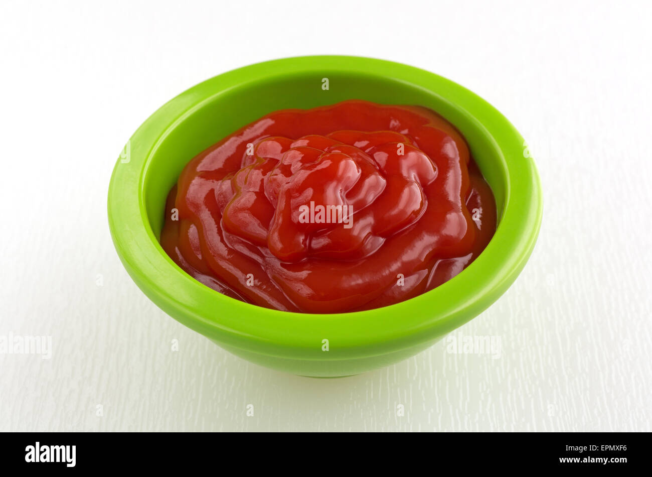 A small green portion bowl filled with ketchup on a white cutting board. Stock Photo