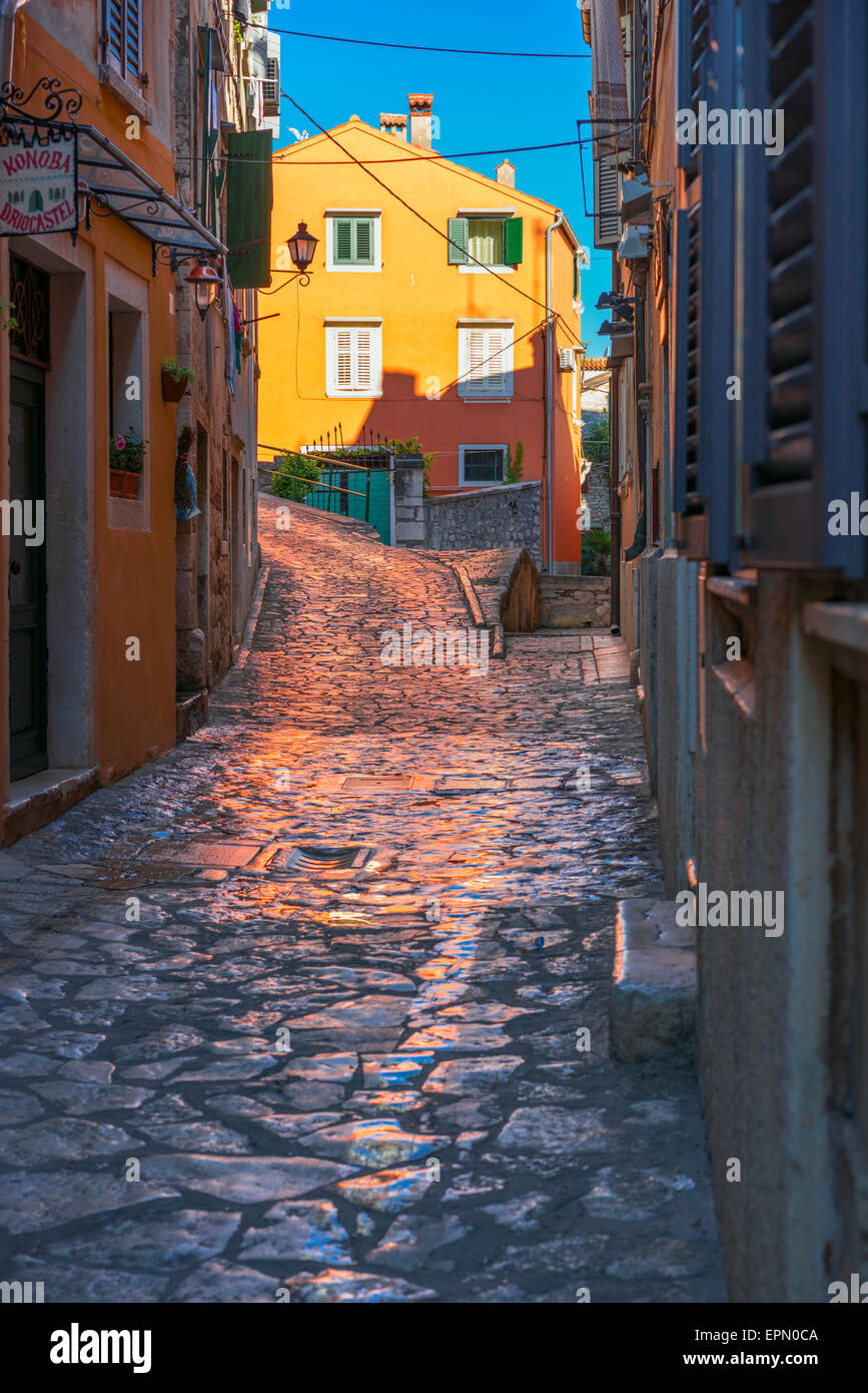 Morning sun shines on an orange building in Rovinj, Croatia, and casts an orange glow brightly on the wet cobble stones below. Stock Photo