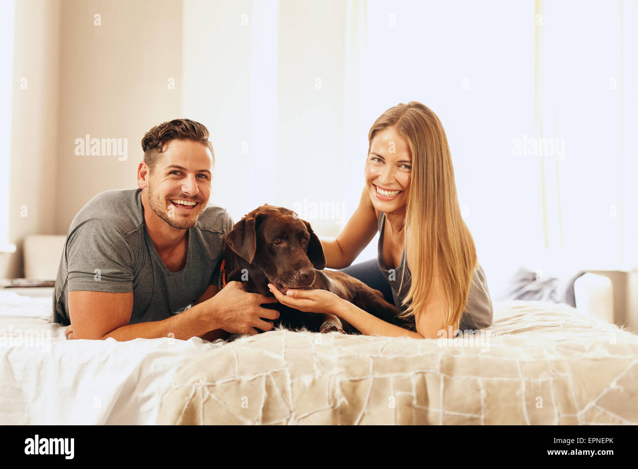 Shot of a young couple lying on the bed with their dog. Cheerful man and woman looking at camera with their pet in bedroom. Stock Photo