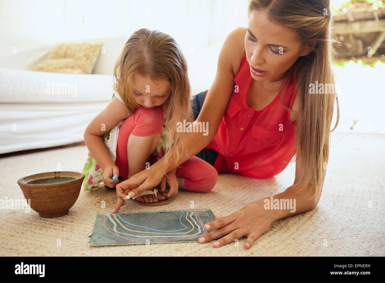 Portrait of mother and little daughter together drawing pictures using color chalks, both sitting on floor in living room at hom Stock Photo
