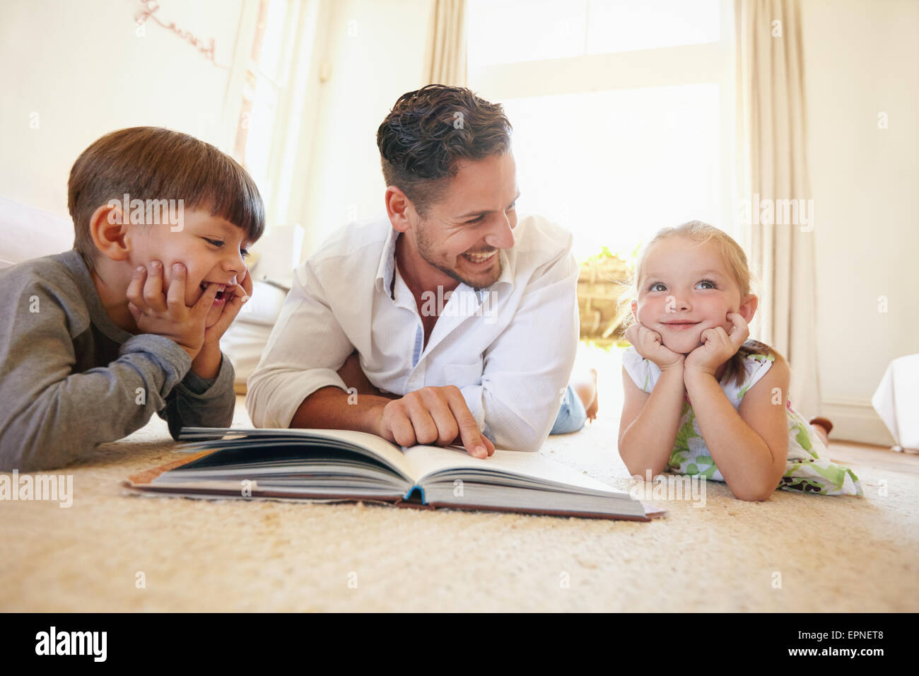 Portrait of happy young family of three lying on floor with a book. Father with two kids reading a story book in living room at Stock Photo