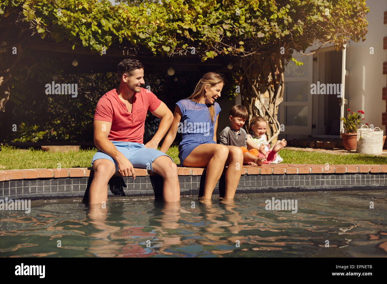 Happy young couple sitting on the edge of swimming pool with their kids enjoying a hot summer day near pool. Couple's feet in wa Stock Photo