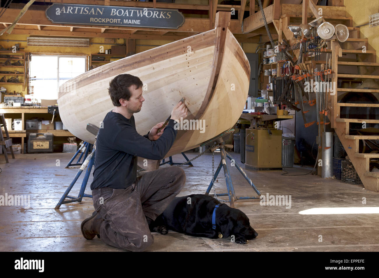 A boat builder,  with his dog at his side, works on the boat he is restoring. Stock Photo