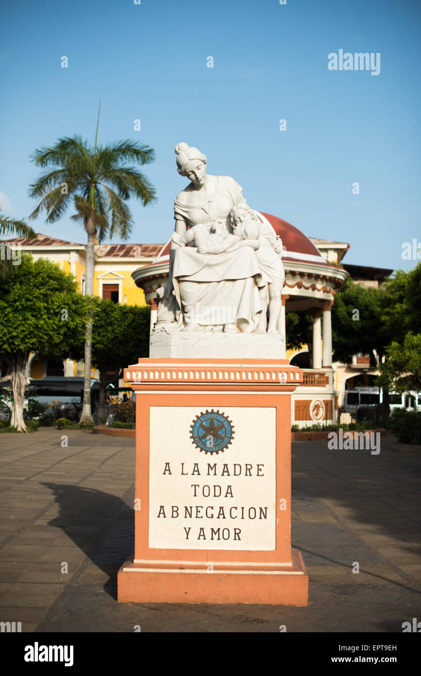 Motherhood Statue Parque Central Granada Nicaragua // GRANADA, Nicaragua — A statue honoring motherhood stands prominently in Parque Central, the main square and historic heart of Granada. The monument features an inscription that reads 'Mother of all devotion and love,' celebrating maternal dedication. Parque Central serves as a central gathering place for locals and visitors in Granada, one of Nicaragua's oldest colonial cities. The square is surrounded by colorful Spanish colonial architecture and important civic buildings. This public artwork represents one of many cultural monuments found Stock Photo
