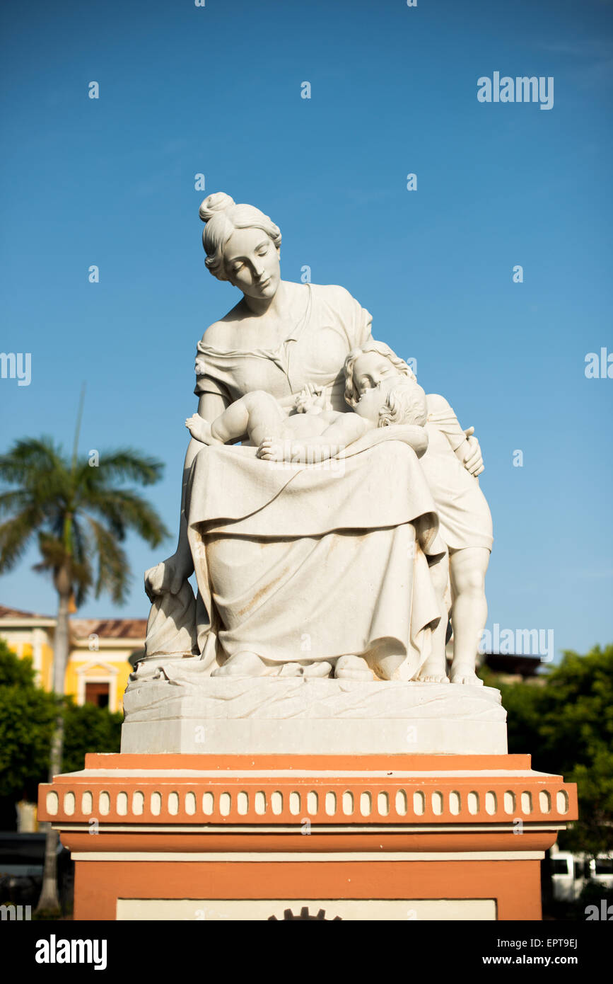 Motherhood Statue Parque Central Granada Nicaragua // GRANADA, Nicaragua — A statue honoring motherhood stands prominently in Parque Central, the main square and historic heart of Granada. The monument features an inscription that reads 'Mother of all devotion and love,' celebrating maternal dedication. Parque Central serves as a central gathering place for locals and visitors in Granada, one of Nicaragua's oldest colonial cities. The square is surrounded by colorful Spanish colonial architecture and important civic buildings. This public artwork represents one of many cultural monuments found Stock Photo