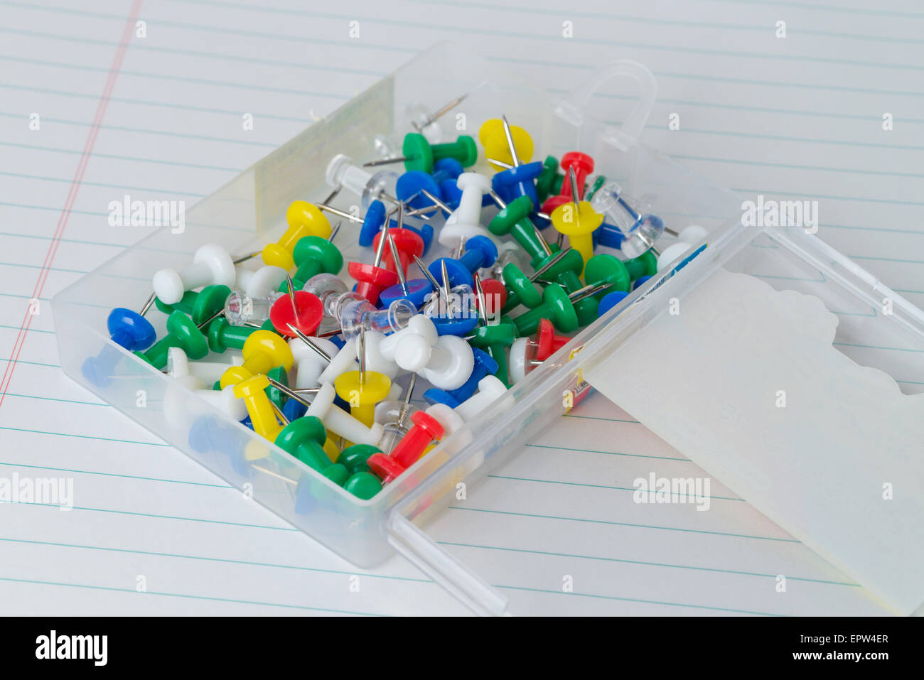 A plastic case full of multicolored thumb tacks on a white piece of lined loose leaf paper. Stock Photo