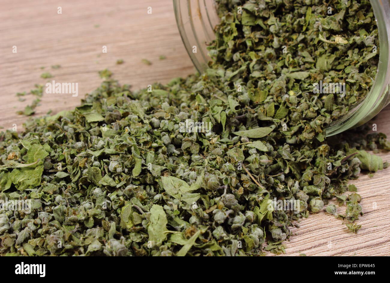 Heap of dried marjoram spilling out of glass jar on wooden background