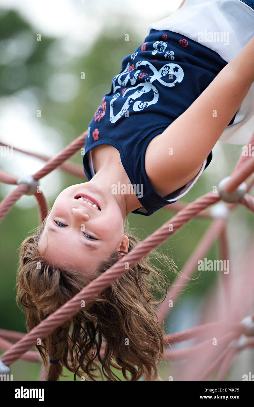 Young girl hangs upside down from a rope outdoor jungle gym at an outdoor playground park Stock Photo