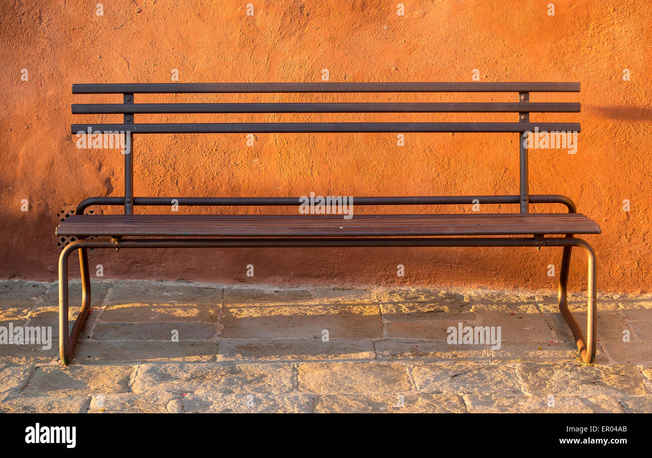 Iron bench against an orange painted wall and a stone floor lit by the evening sun Stock Photo