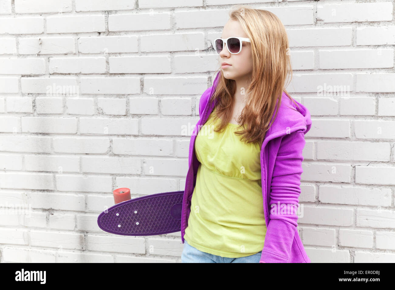 Blond teenage girl in sunglasses holds skateboard near white urban brick wall Stock Photo