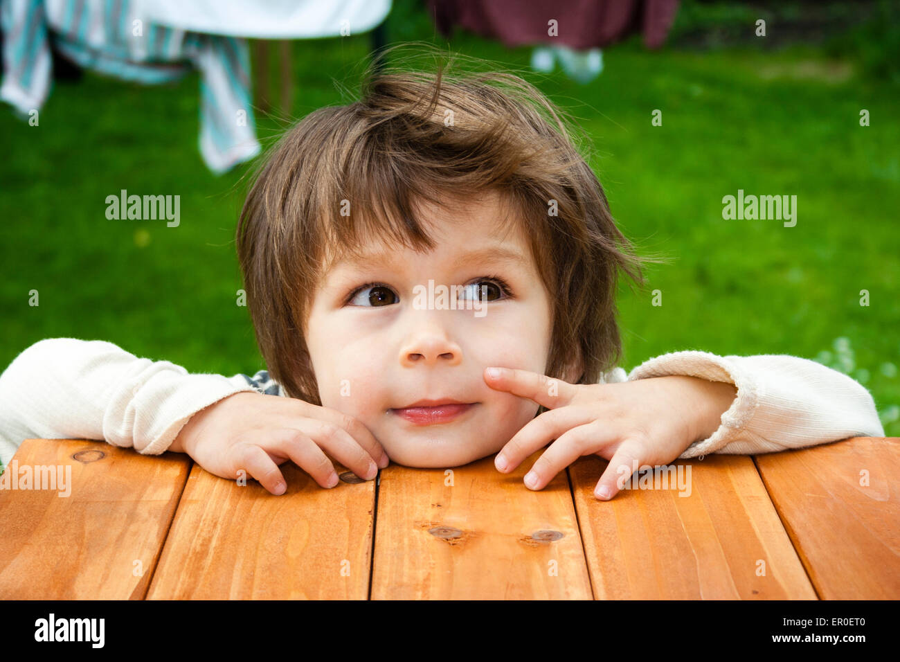 Close up of young child's face, boy, 3-4 year old, as he rests chin and hands on wooden platform, facing and looking to the side. Outdoors Stock Photo