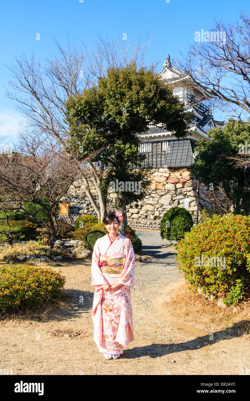 Young Japanese women standing posing for viewer while wearing a pink kimono and a flower in her hair outdoors with Hamamatsu castle in background. Stock Photo