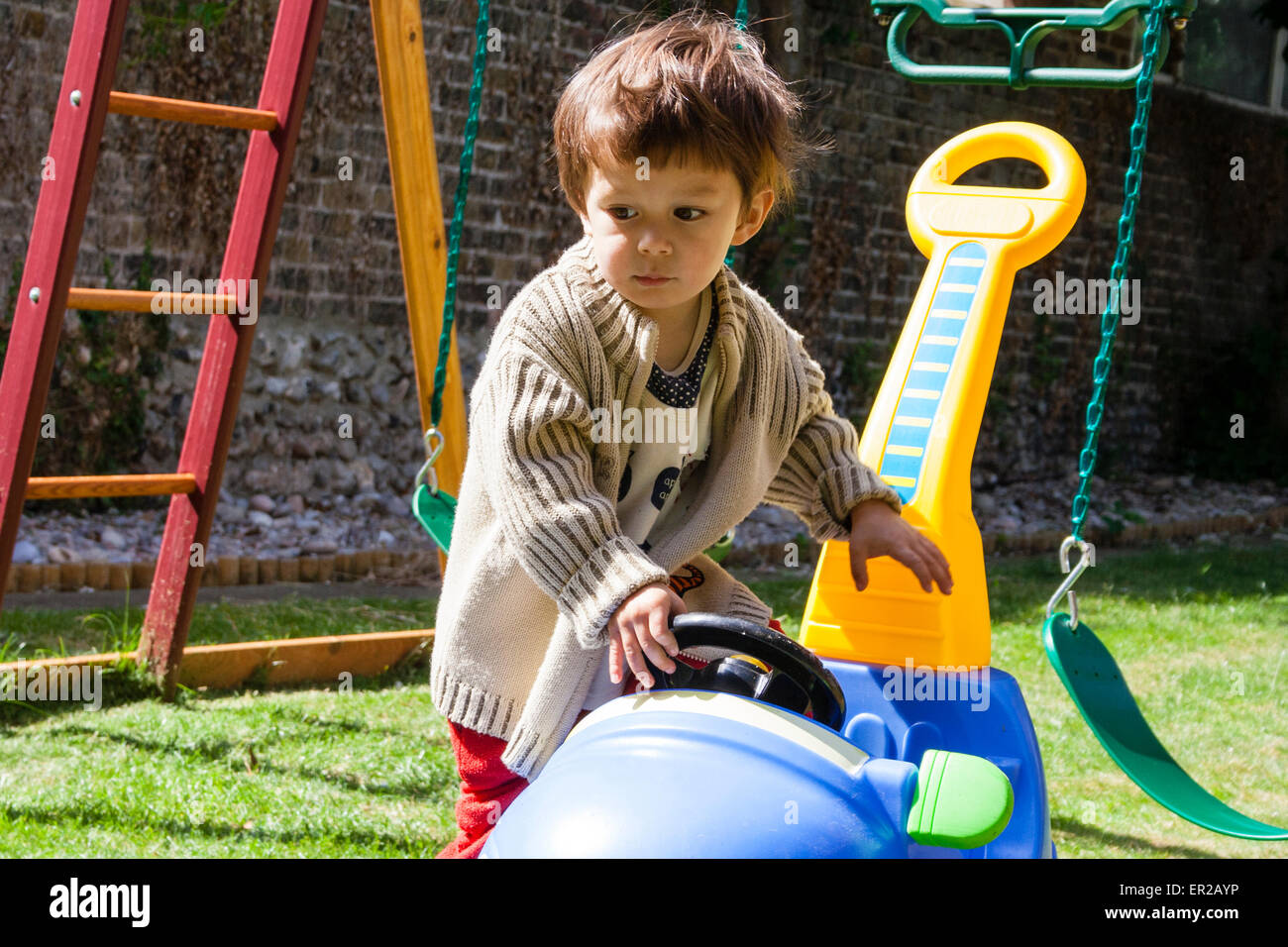Child, boy, 4-5 year old, wearing a cardigan and playing by pushing around a blue and yellow plastic sit in car in a garden on a sunny day. Stock Photo