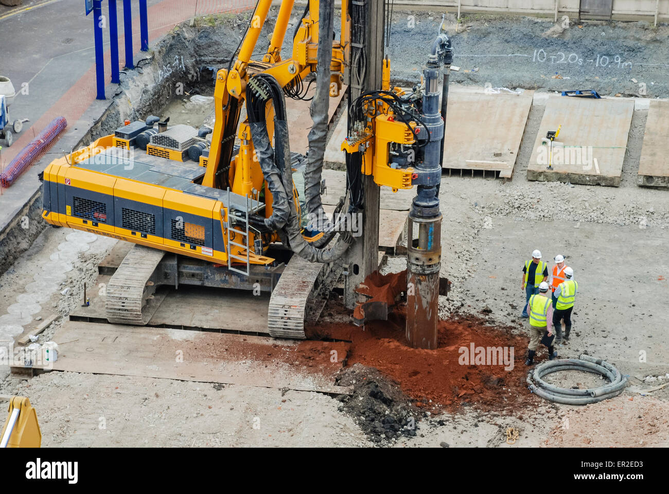 Four builders stand beside a large machine as it inserts a metal pile into the ground. Stock Photo